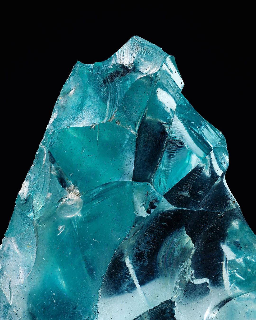 Close-up studio shot of a teal blue gemstone-like mineral with sharp edges against a black background, showcasing its transparency and structure.