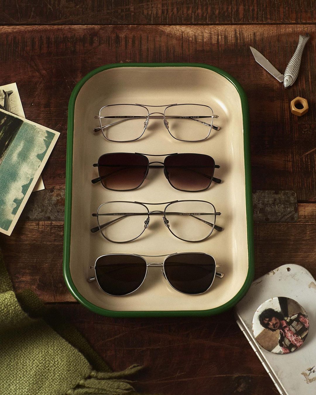 Overhead shot of four pairs of sunglasses arranged in an enamel tray, on a rustic wood surface with vintage postcards and accessories.