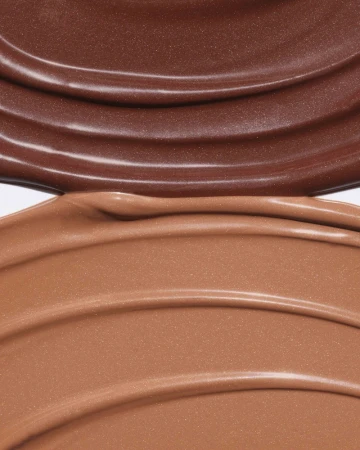 Two shades of glossy, brown liquid foundation or makeup swatched in thick, curving lines on a smooth, solid surface.