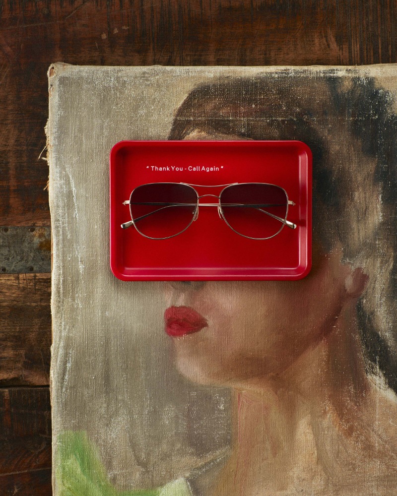 Aviator sunglasses rest on a red tray atop a painterly female portrait; composition balances product display with artistic backdrop.