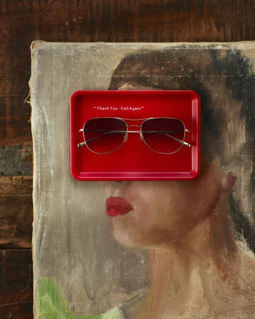 Aviator sunglasses rest on a red tray atop a painterly female portrait; composition balances product display with artistic backdrop.