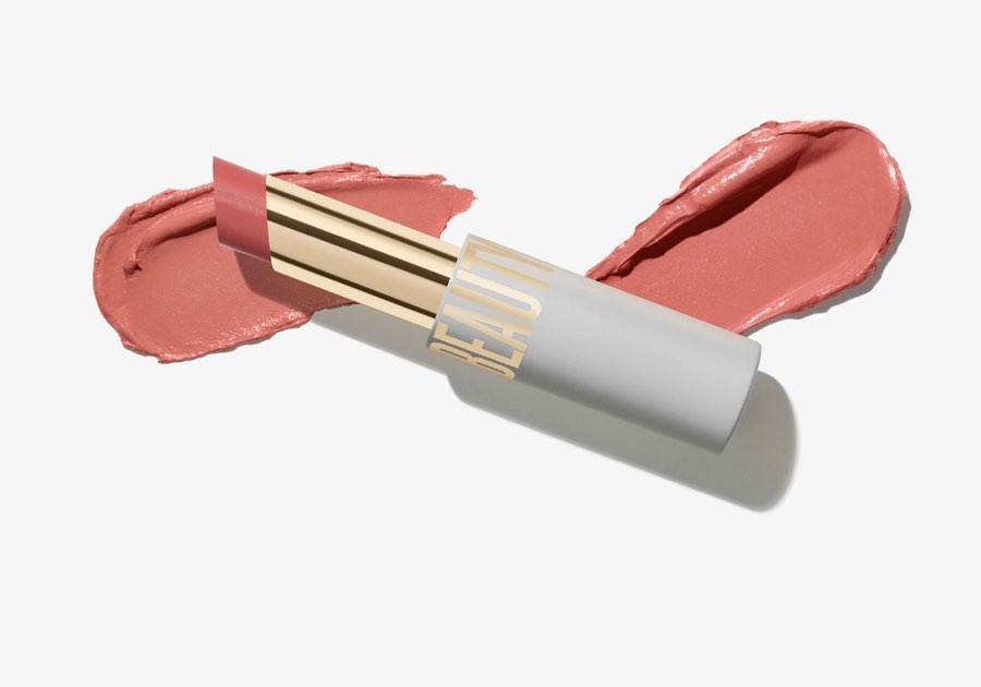 Close-up shot of lipstick with a gold band and pale grey tube lying on a pale pink swatch, all on a white background. Beauty shot.