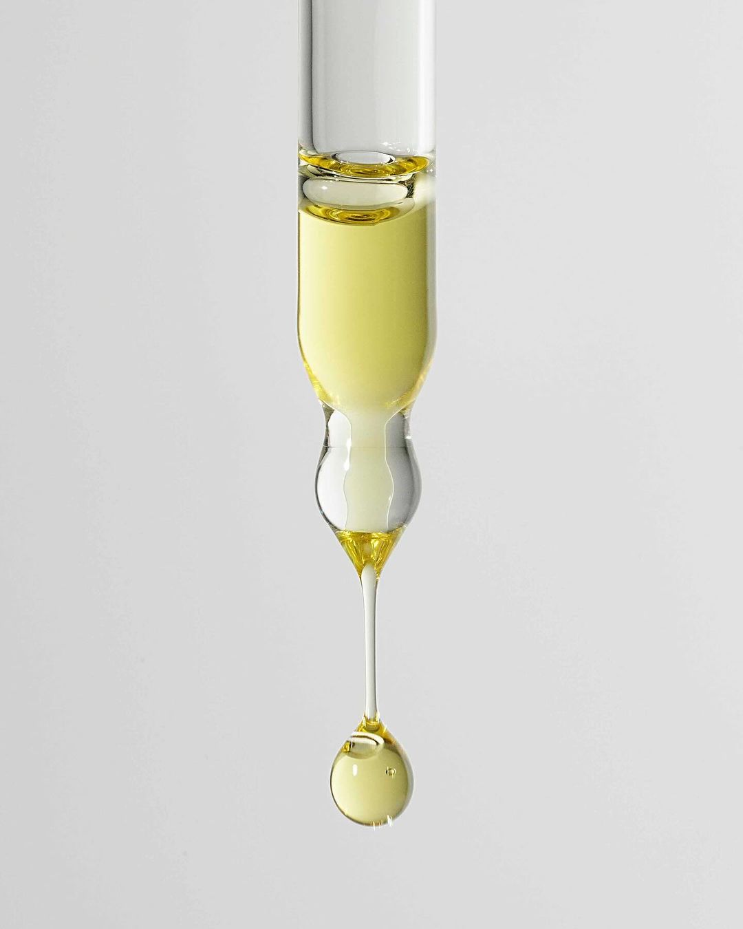 Close-up of a glass dropper with a droplet of light yellow liquid, likely cosmetic oil or serum, against a neutral gray background.