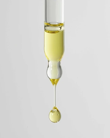 Close-up of a glass dropper with a droplet of light yellow liquid, likely cosmetic oil or serum, against a neutral gray background.