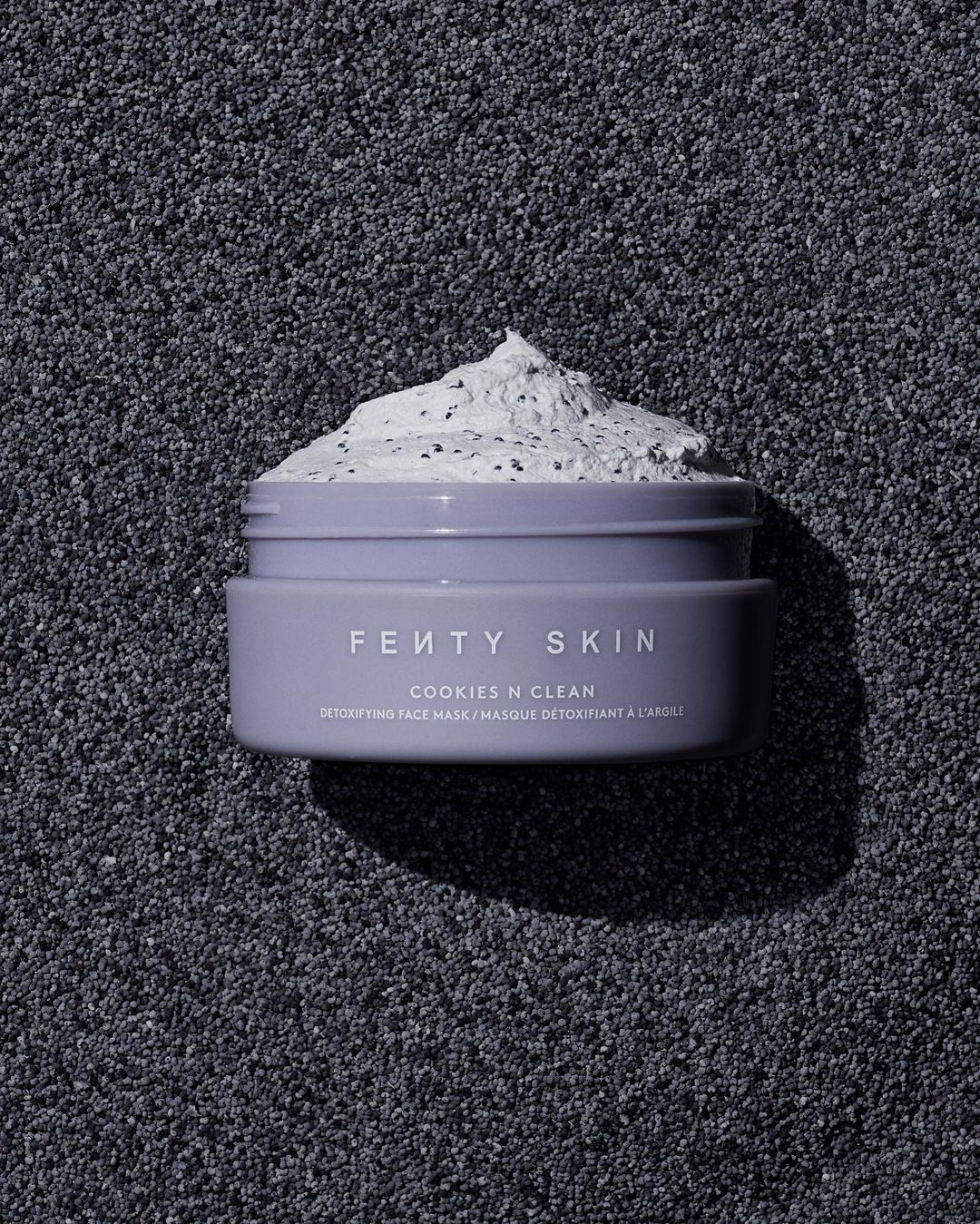Close-up of Fenty Skin Cookies N Clean detoxifying face mask in a lavender jar on a textured dark grey background.