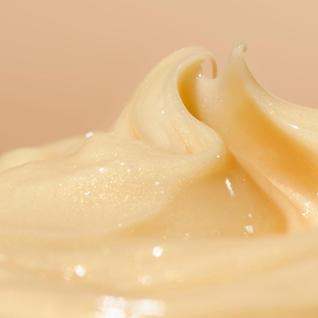 Close-up of pale-yellow cream swirled in a peak, glistening in the light against a soft cream background; beauty product advertising.