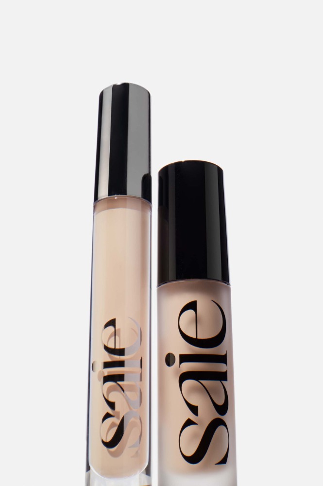 Two bottles of Saie makeup are side-by-side against a white background, one with a silver cap and the other with a black cap.