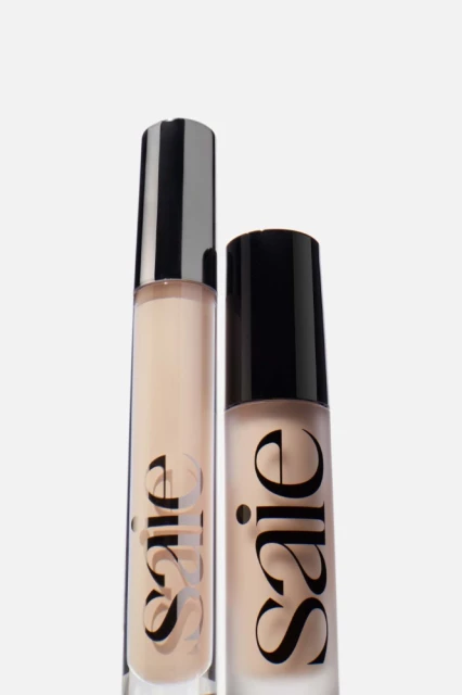 Two bottles of Saie makeup are side-by-side against a white background, one with a silver cap and the other with a black cap.