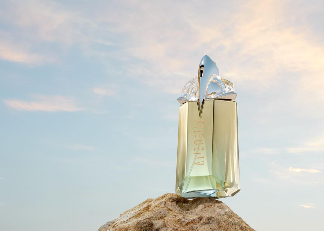 Faceted glass perfume bottle resting upright on a weathered rock against a soft sunset sky backdrop.