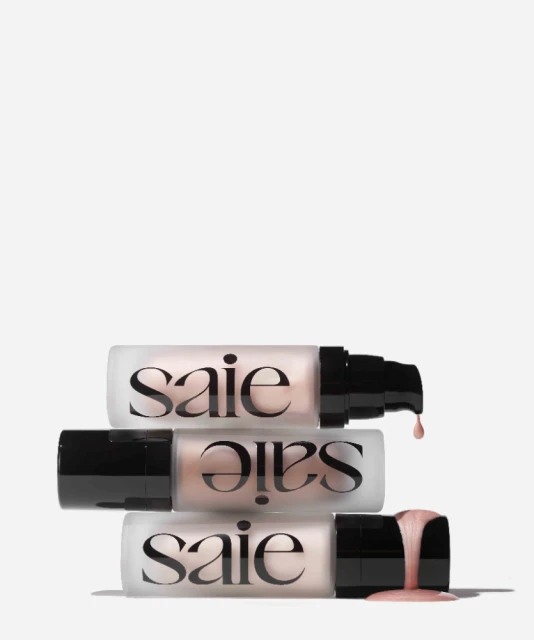Three bottles of Saie brand illuminating serum stacked on a white background with product dripping from the applicator of the top bottle.