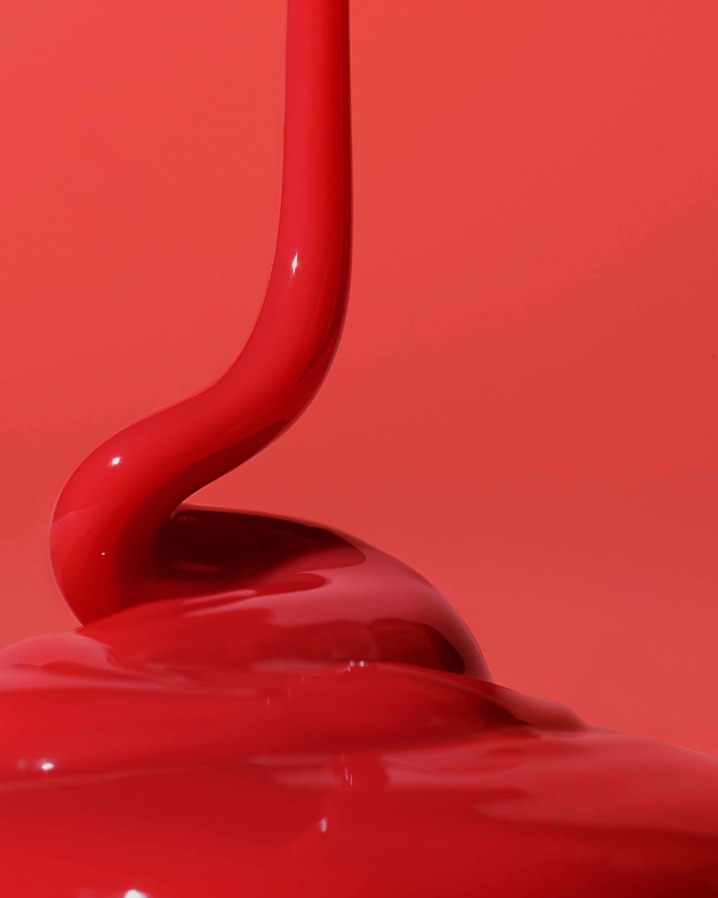 Bold, shiny red liquid is captured in a close-up studio shot with a vibrant, monochromatic red backdrop, showcasing texture and movement.