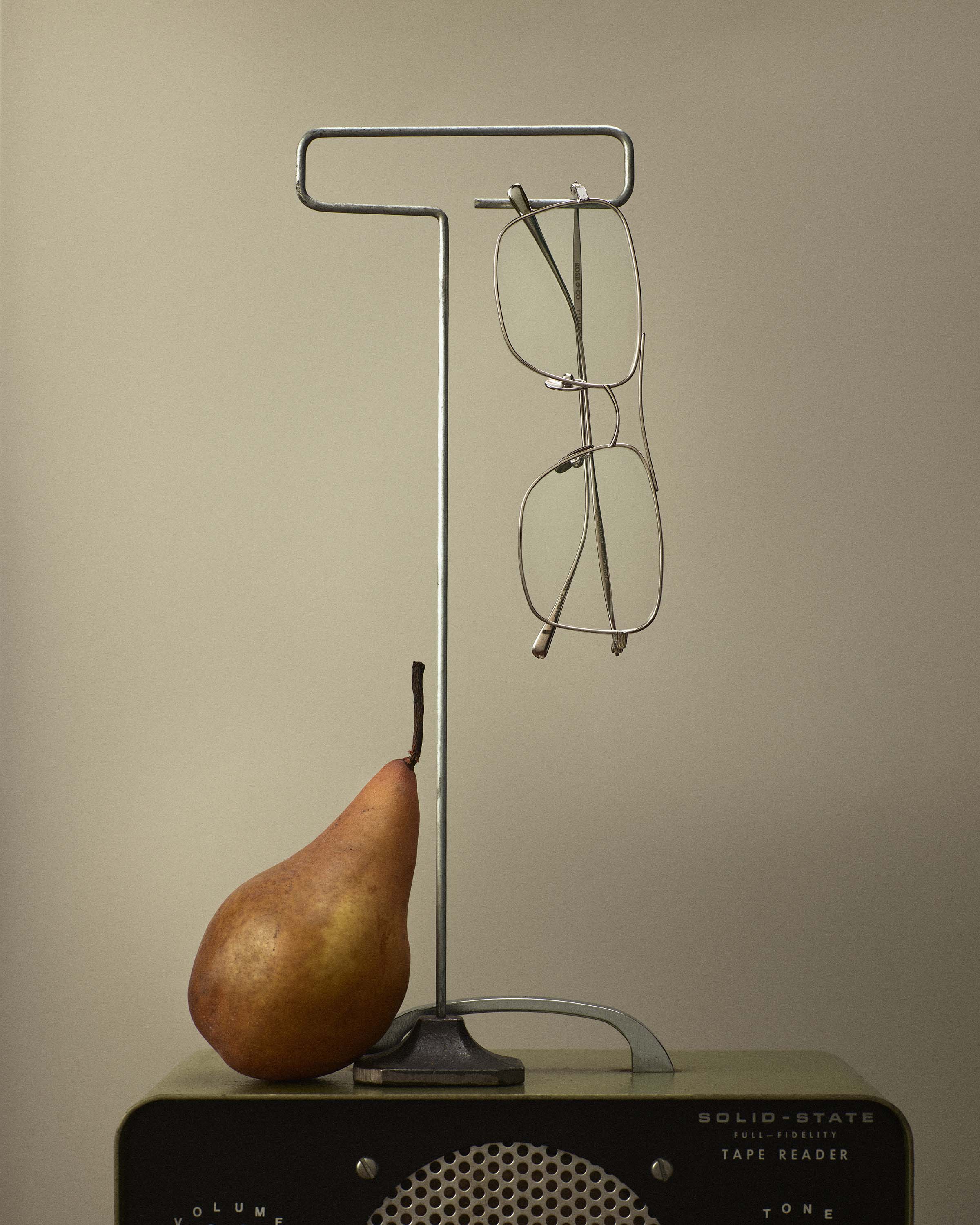 Still life composition featuring eyeglasses hanging from a metal stand, a pear, and a vintage radio on a beige background.