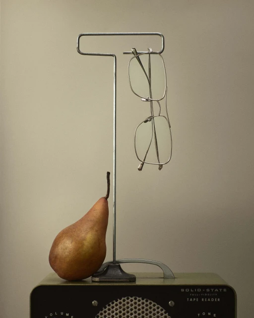 Still life composition featuring eyeglasses hanging from a metal stand, a pear, and a vintage radio on a beige background.