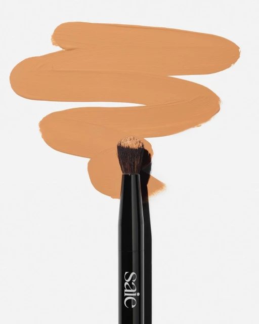 Close-up of a makeup brush with black handle, next to a smear of tan foundation against a white backdrop, beauty photography.