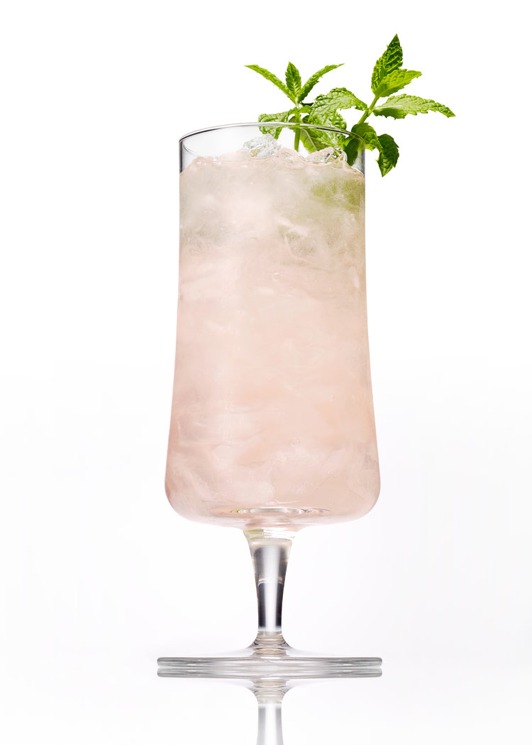 Tall glass of a refreshing pink cocktail with crushed ice and a sprig of green mint on a white background in a studio setting.
