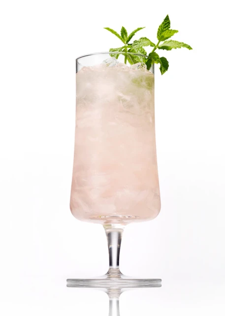 Tall glass of a refreshing pink cocktail with crushed ice and a sprig of green mint on a white background in a studio setting.