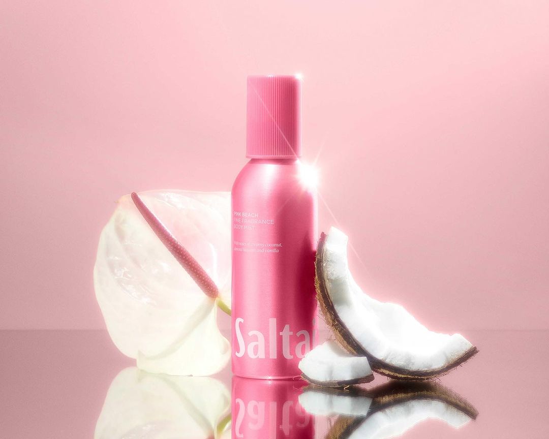 Pink spray bottle of Saltair body treatment with coconut and anthurium flower on a reflective surface against a pink background.