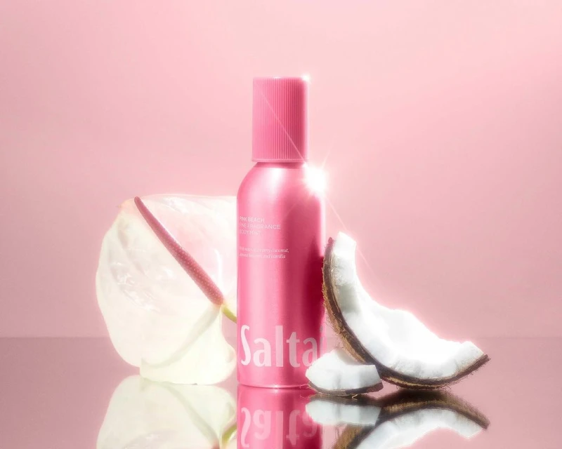 Pink spray bottle of Saltair body treatment with coconut and anthurium flower on a reflective surface against a pink background.