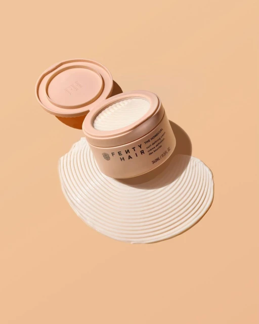 Cosmetic pot from Fenty Hair sitting on swirling creme on peach background in studio shot for beauty and hair care advertising.
