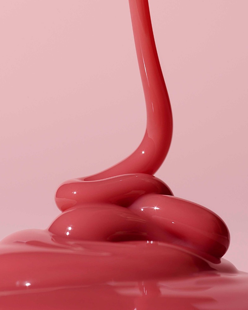 Close-up shot of thick, glossy pink fluid dripping and pooling against a pale pink backdrop, highlighting texture and movement.