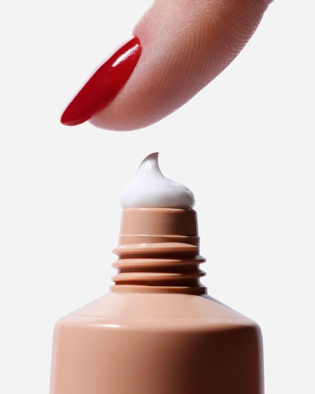 Close-up of fingertip with long red nail polish pressing cream onto a tan tube against a white background in a beauty studio shot.
