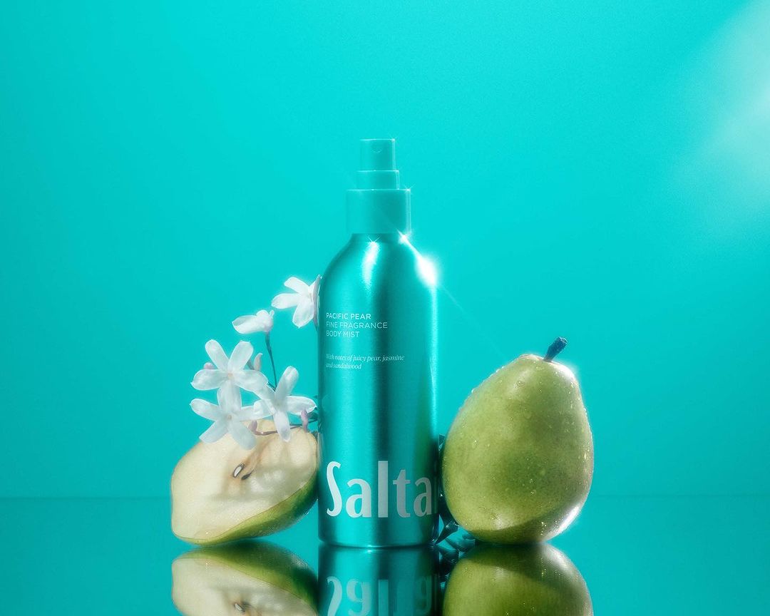 Turquoise fragrance body mist product shot with whole and sliced pear, and white flowers, on reflective surface for beauty advertising.