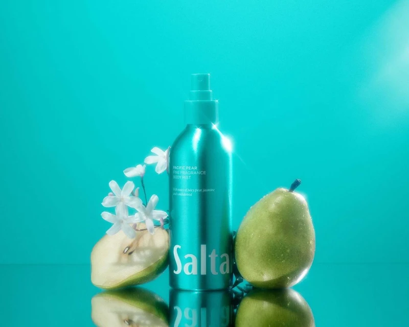 Turquoise fragrance body mist product shot with whole and sliced pear, and white flowers, on reflective surface for beauty advertising.