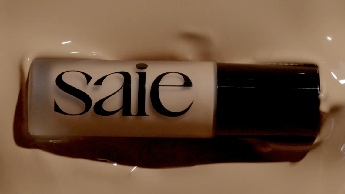 Elevated view of a bottle of Saie foundation surrounded by liquid foundation makeup product, promoting beauty and cosmetic brands.