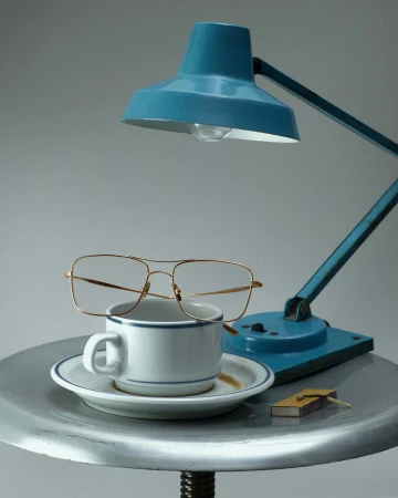 A vintage-style still life photograph featuring a teal desk lamp, glasses, a white cup and saucer, and a matchbox on a metal table.