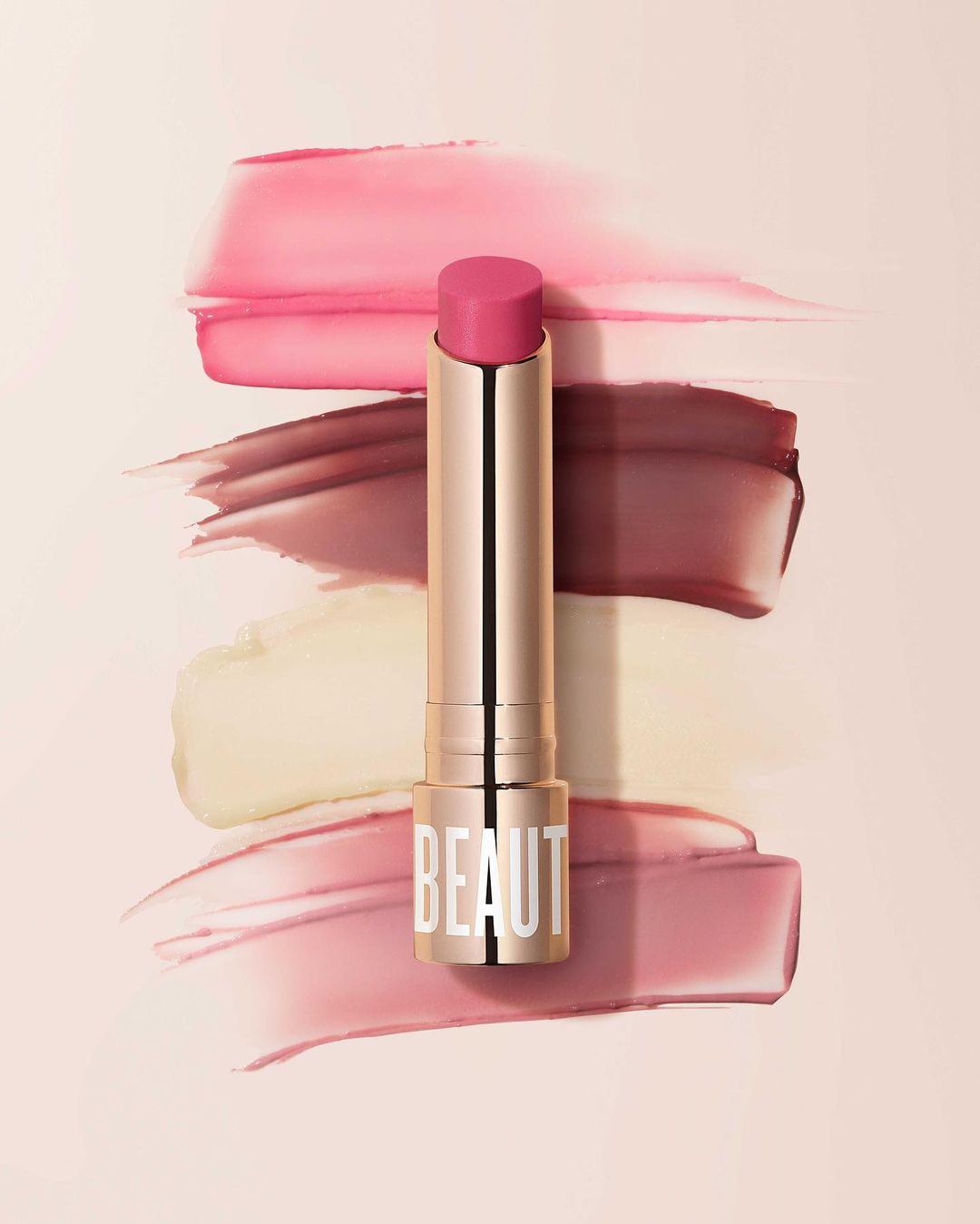 Still-life product shot of a pink lipstick tube with the text 'BEAUT' against streaks of various makeup product colours in the background.