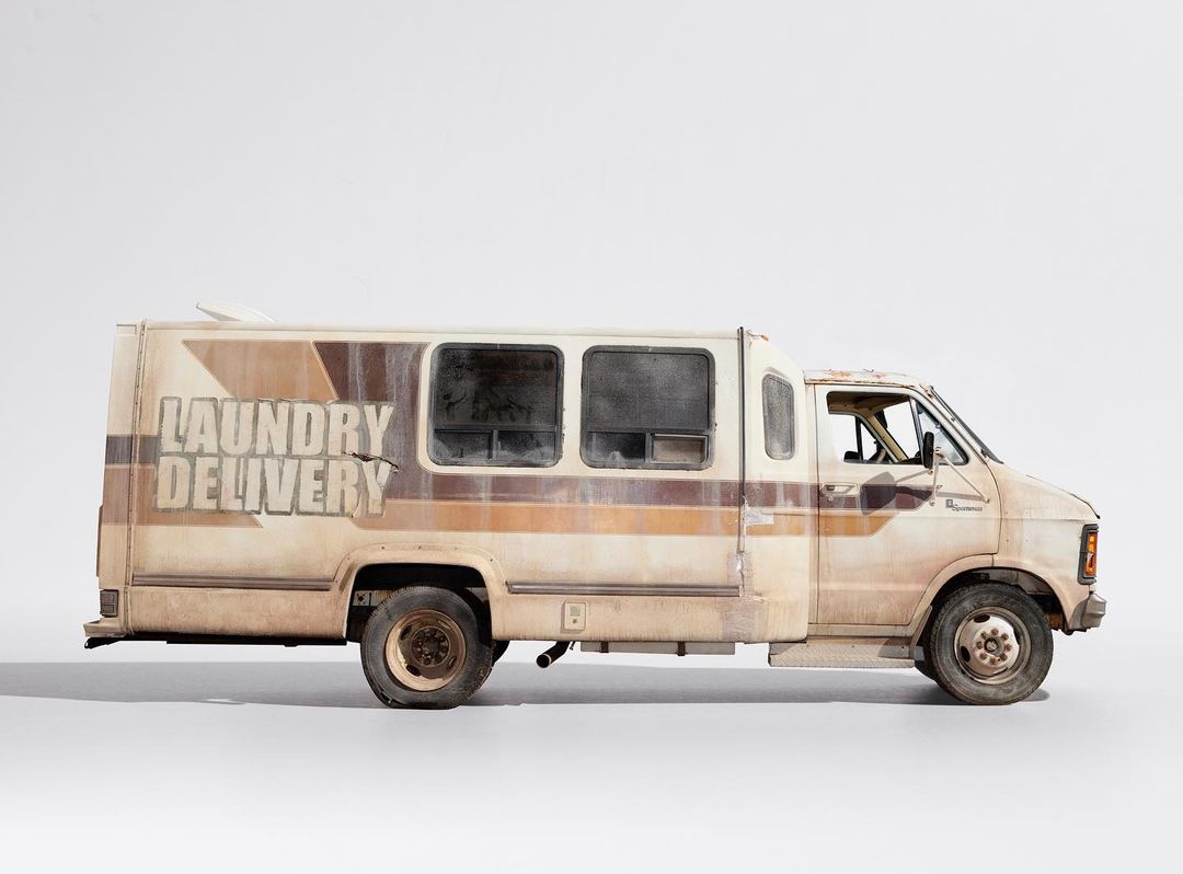 Profile shot of a weathered, cream-colored laundry delivery van parked in a white studio, showcasing its textured exterior and unique design.