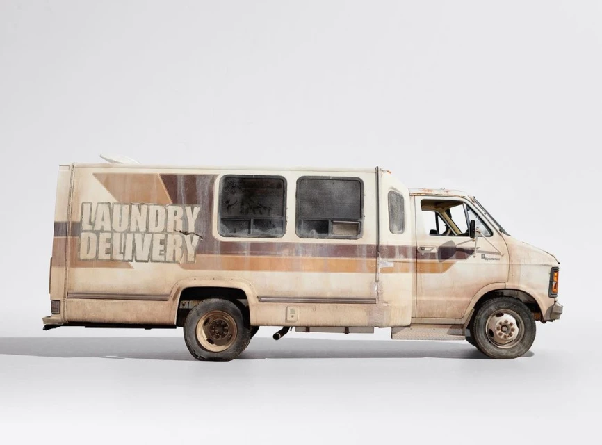 Profile shot of a weathered, cream-colored laundry delivery van parked in a white studio, showcasing its textured exterior and unique design.