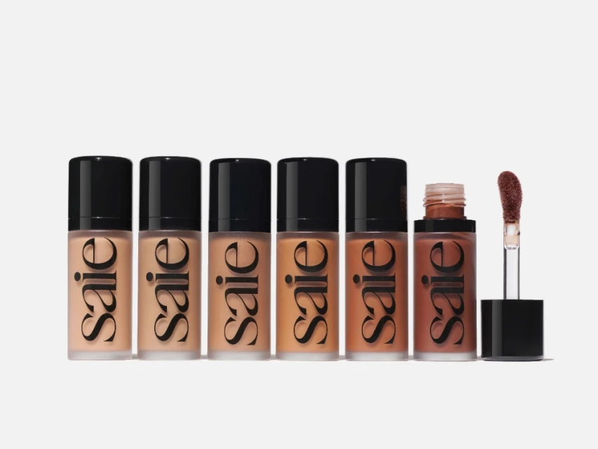 Six 'Saie' brand foundation bottles in various shades, aligned on a white background. Makeup brush applicator positioned next to the last bottle.