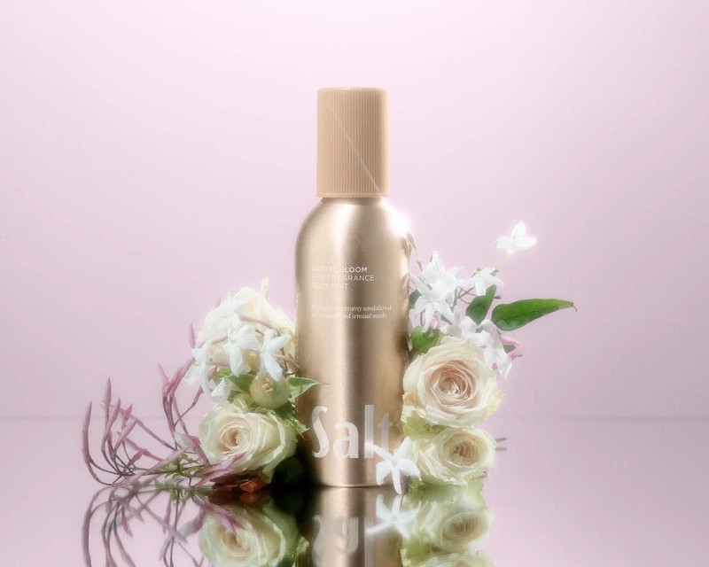 Product shot of a gold spray bottle surrounded by roses and jasmine flowers on a reflective surface with a pink backdrop.
