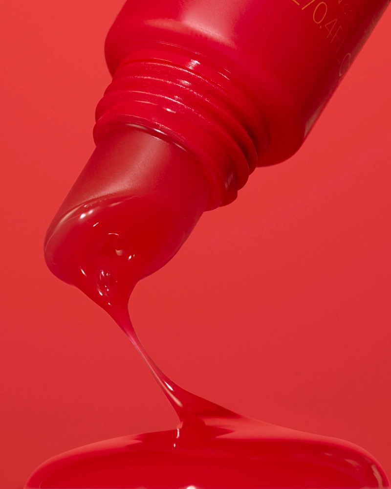 Close-up studio shot of bright red viscous liquid pouring from a red tube onto a red surface in a monochrome setting.