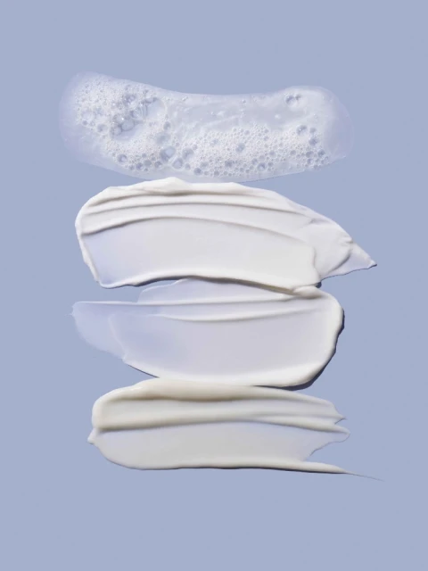 Close-up of cosmetic textures arranged on a purple background, including clear cleanser with bubbles and three shades of cream.