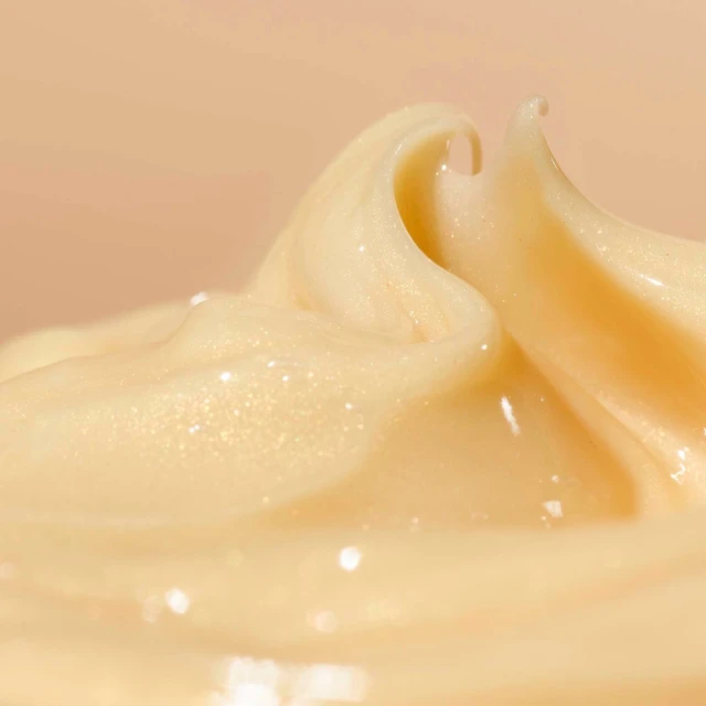 Macro shot of creamy, pale yellow beauty product, likely skincare or haircare, with a smooth, glossy texture against a soft peach backdrop.