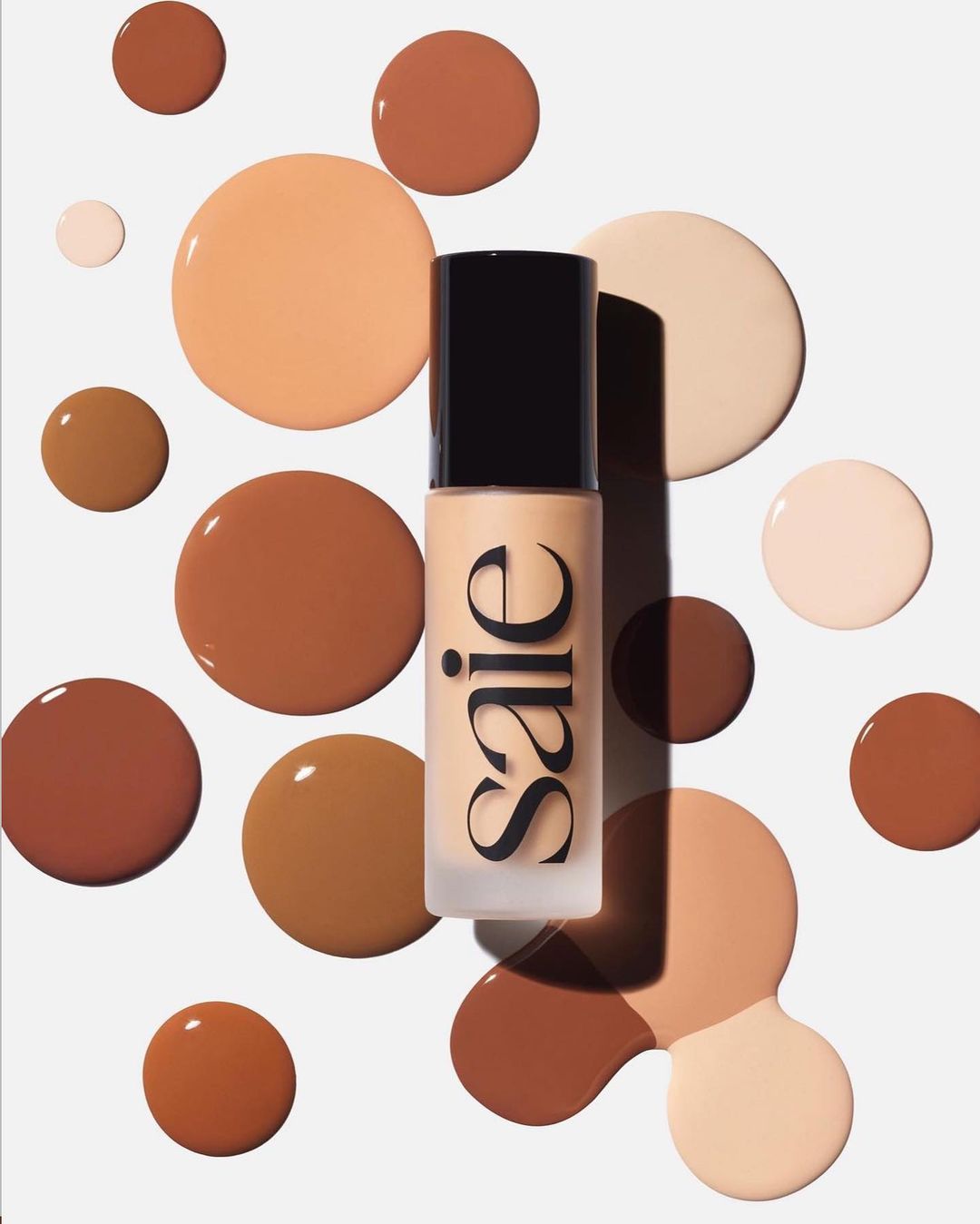 Cosmetic bottle with Saie logo surrounded by brown, beige, and tan liquid foundation splatters on a solid white backdrop for beauty content.