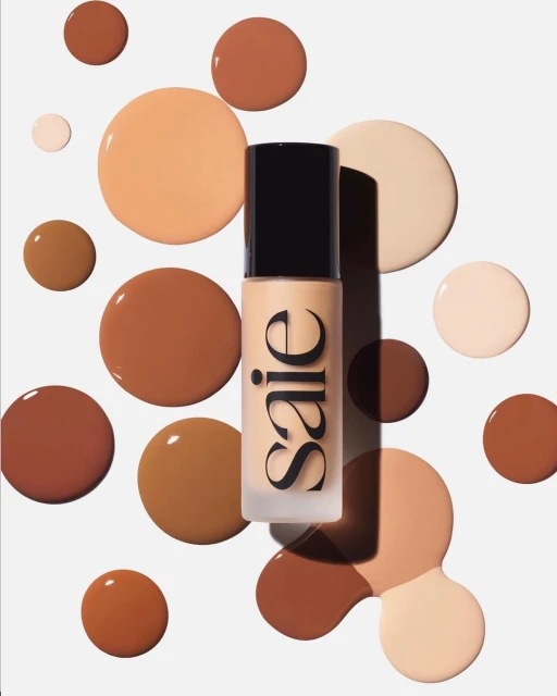 Cosmetic bottle with Saie logo surrounded by brown, beige, and tan liquid foundation splatters on a solid white backdrop for beauty content.