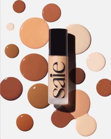 Cosmetic bottle with Saie logo surrounded by brown, beige, and tan liquid foundation splatters on a solid white backdrop for beauty content.