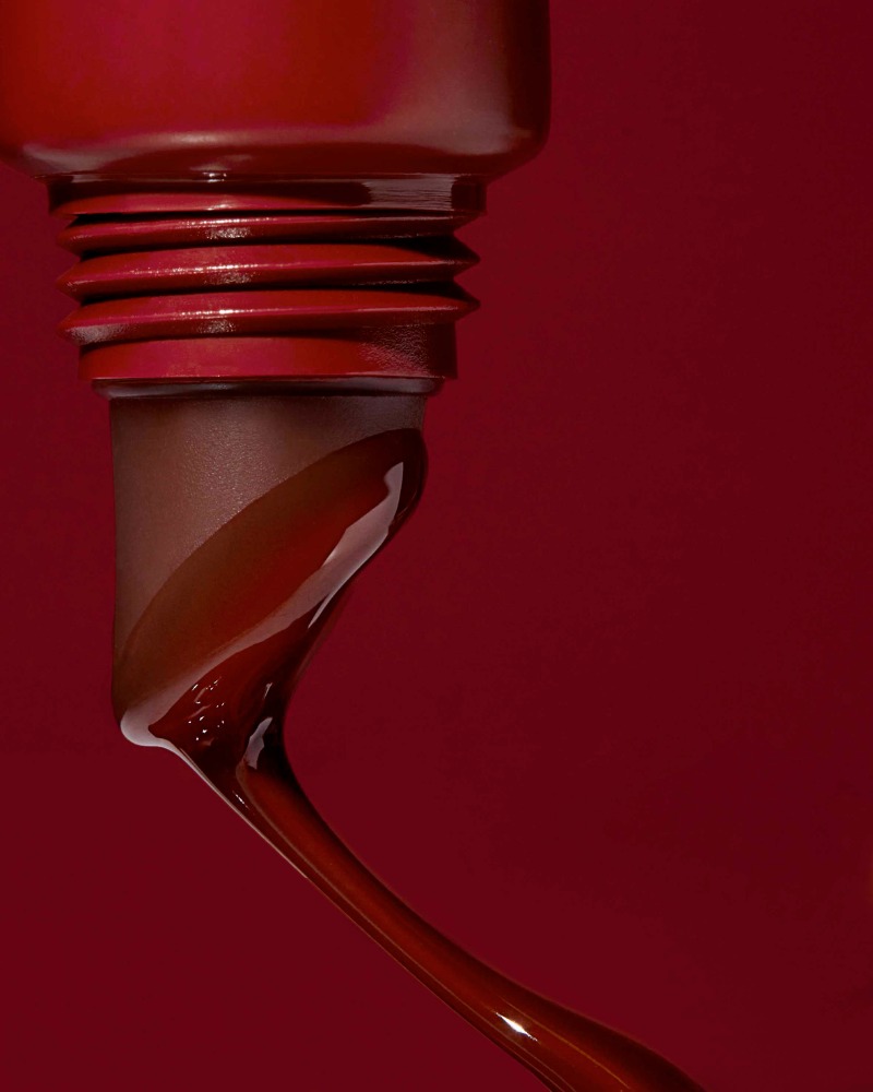 Close-up of a dark red, glossy liquid flowing from a container, set against a matching dark red background for beauty and fashion.