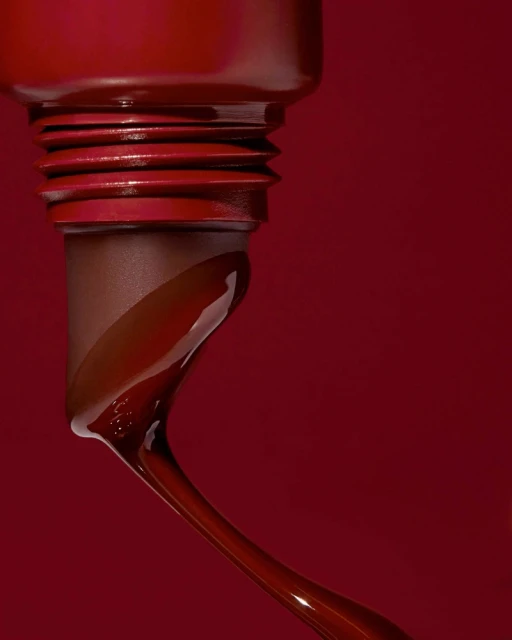 Close-up of a dark red, glossy liquid flowing from a container, set against a matching dark red background for beauty and fashion.