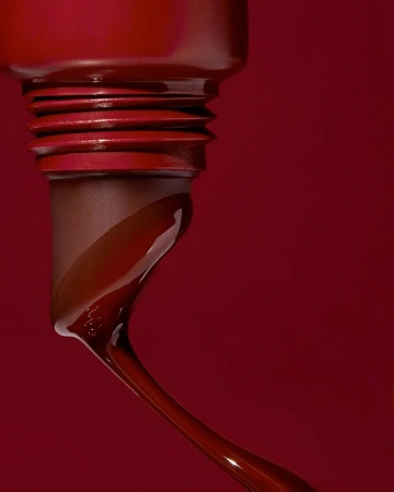 Close-up of a dark red, glossy liquid flowing from a container, set against a matching dark red background for beauty and fashion.