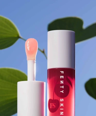 Fenty Skin cherry lip oil with wand applicator, featuring white packaging and green foliage against a bright blue sky backdrop.