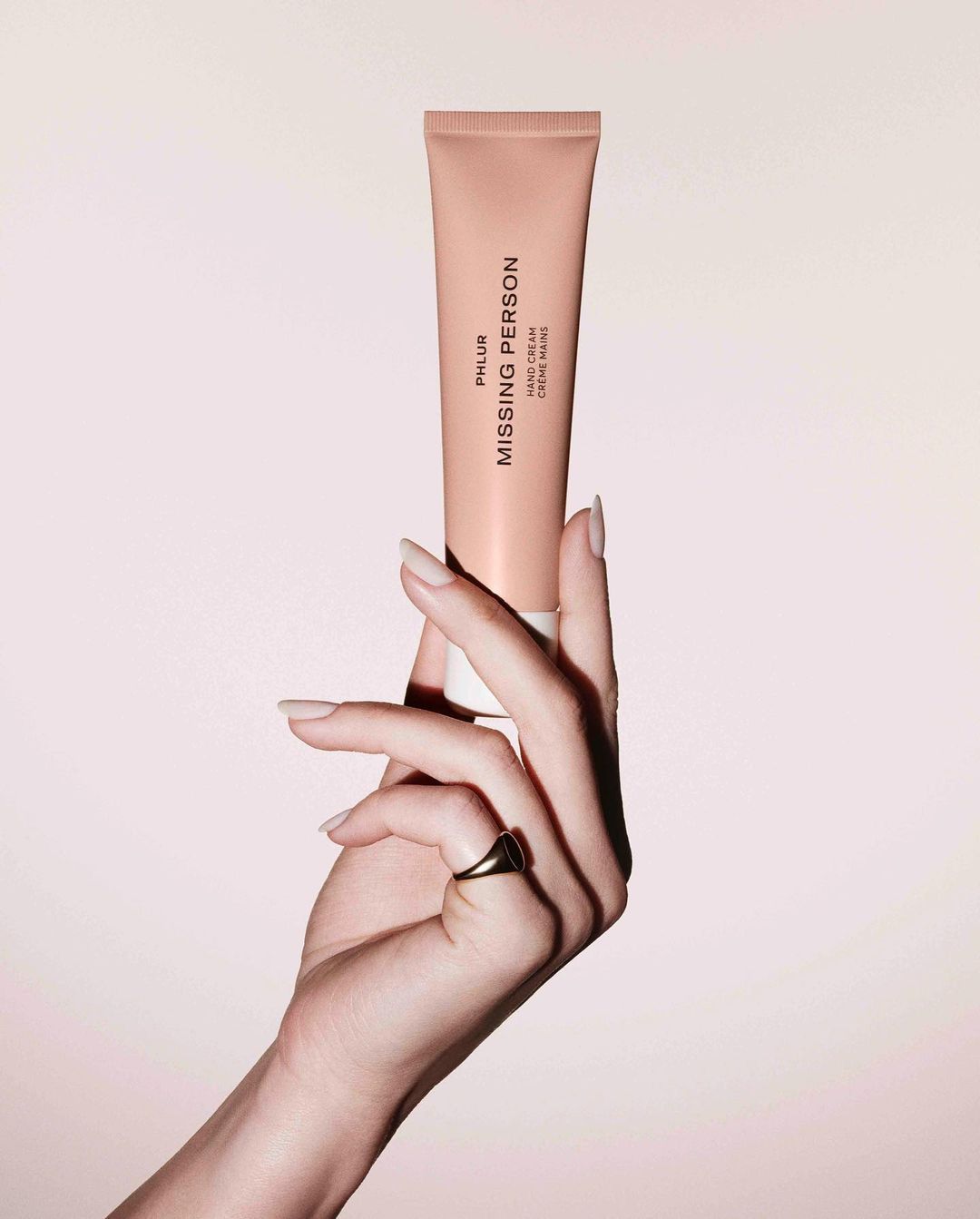 A hand holds a Phlur Missing Person hand cream tube against a soft pink backdrop. The product and manicured hand are in focus.