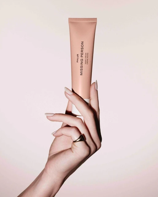 A hand holds a Phlur Missing Person hand cream tube against a soft pink backdrop. The product and manicured hand are in focus.