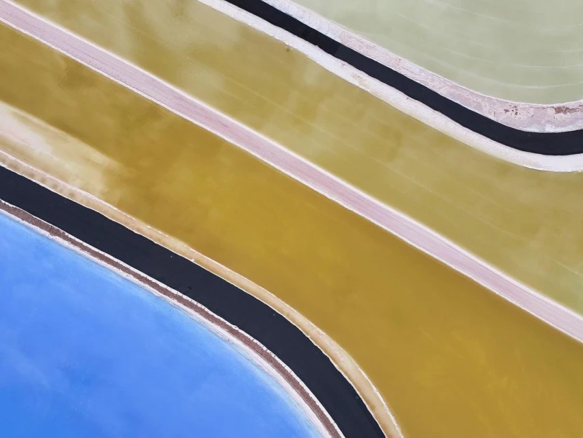 Aerial view of a landscape with colorful pools of blue, green, and yellow water separated by narrow black channels and sandy ground.