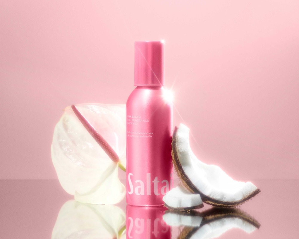 Pink Salt Air bottle sits alongside a white anthurium flower and slices of coconut on a reflective surface, against a pink backdrop.