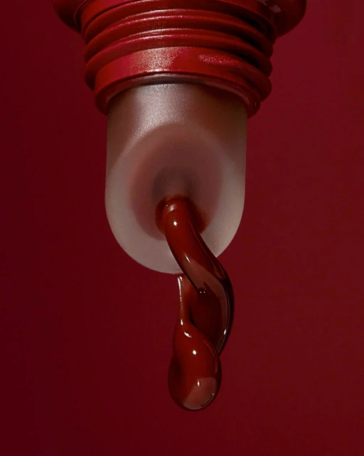 Close-up of viscous, dark red liquid dripping from translucent cosmetic container against a solid red background in studio lighting.