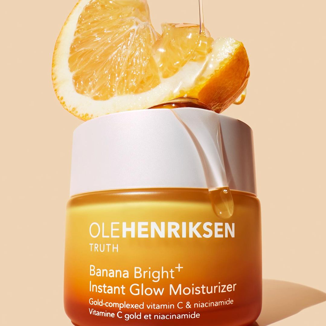 Ole Henriksen Banana Bright+ Instant Glow Moisturizer jar with orange slices and dripping liquid on a peach-colored background.